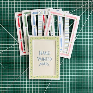 May include: A collection of rectangular white paper mats featuring hand-painted borders in various colors including red, green, blue, and pink. The front-most mat displays the text "HAND PAINTED MATS" in blue ink, centered within a light green dotted border.