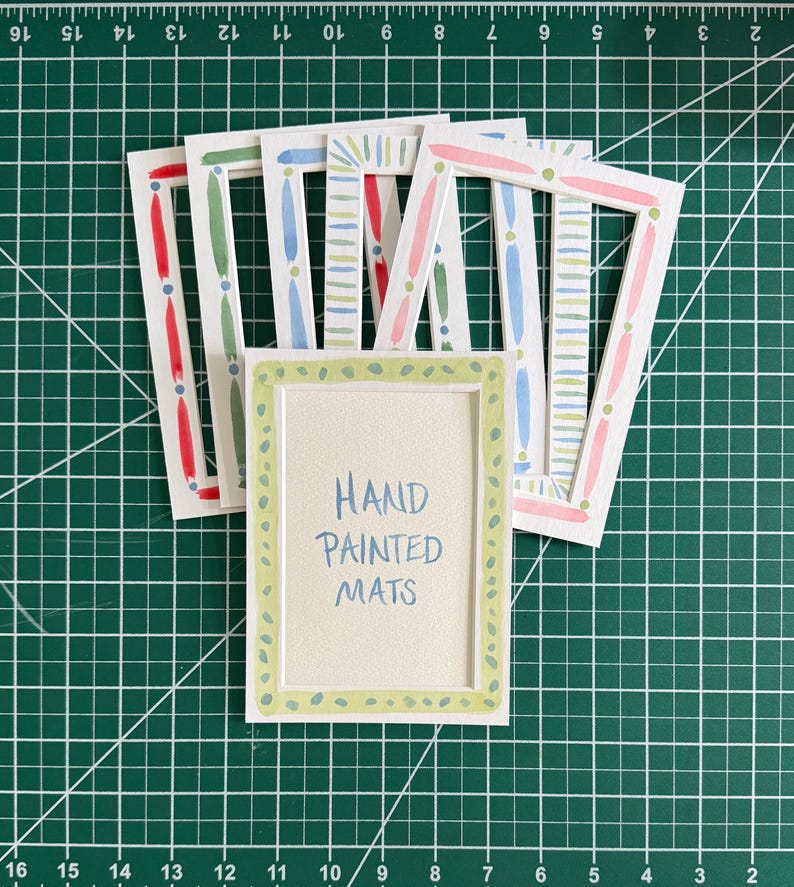 May include: A collection of rectangular white paper mats featuring hand-painted borders in various colors including red, green, blue, and pink. The front-most mat displays the text "HAND PAINTED MATS" in blue ink, centered within a light green dotted border.