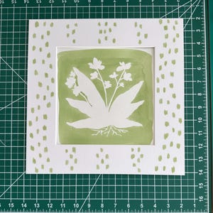 May include: A square white paper mat featuring a sage green painted background with a white silhouette of a plant with roots. The mat has a border of sage green dash marks. The mat measures 15 cm by 15 cm on a green cutting mat with grid lines.
