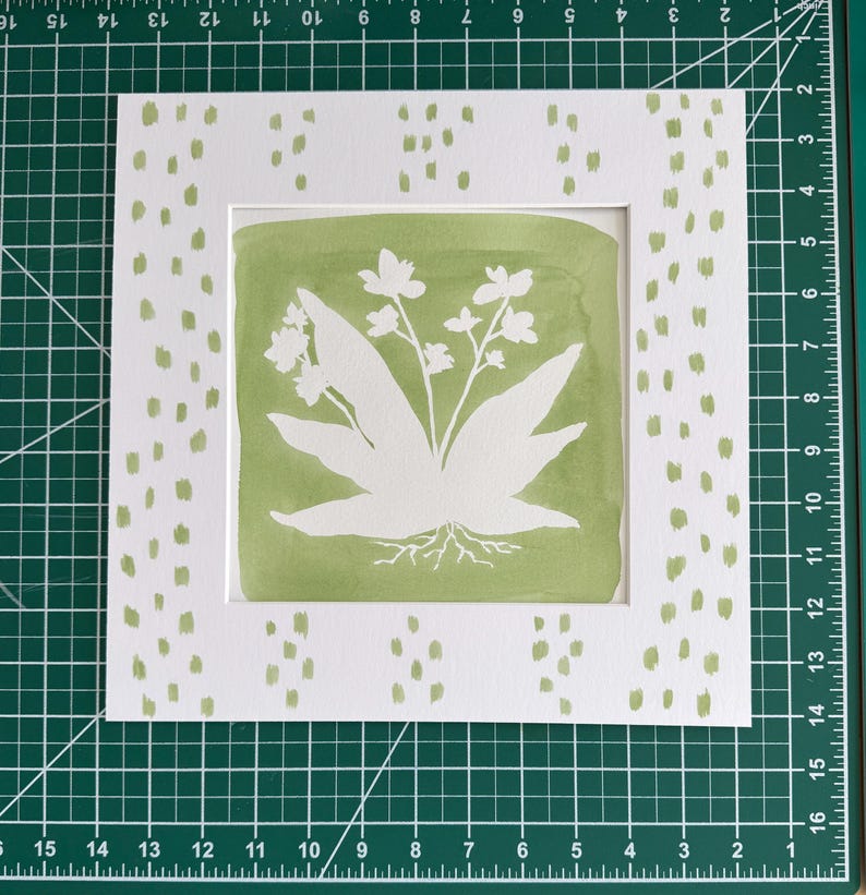 May include: A square white paper mat featuring a sage green painted background with a white silhouette of a plant with roots. The mat has a border of sage green dash marks. The mat measures 15 cm by 15 cm on a green cutting mat with grid lines.