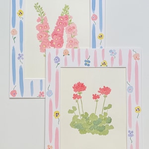 May include: Two rectangular art prints featuring floral illustrations. The top print displays pink hollyhocks, while the bottom print shows red geraniums. Both prints are framed by white borders decorated with vertical brushstrokes and small flowers.