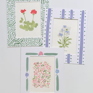 May include: Three rectangular art prints with decorative borders. One features red geraniums in a green wavy-patterned frame. Another shows blue and white pansies in a purple striped frame with dots. The third displays pink flowers in a green frame.
