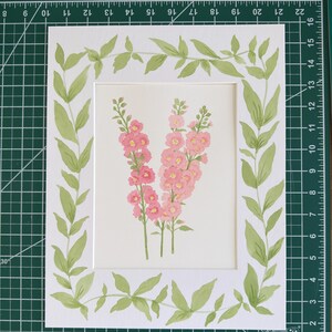 May include: A white paper mat featuring a hand-painted illustration of three pink hollyhock flower stalks in the center, surrounded by a painted border of green leaves and vines. The mat is positioned on a green cutting mat with white grid lines.