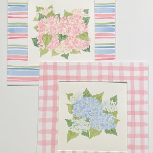 May include: Two square watercolor paintings featuring floral arrangements. The top painting shows pink hydrangea blossoms with green leaves, framed by a border of horizontal blue, pink, and green stripes. The bottom painting displays blue and white hydrangea flowers with green leaves, set within a pink and white checkered border.