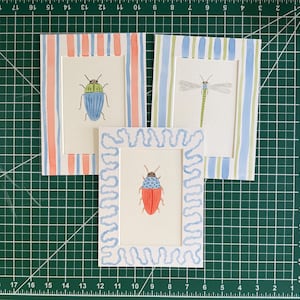 May include: Three rectangular art prints featuring insect illustrations. One print shows a blue beetle in a coral and blue striped frame. Another shows a green dragonfly in a blue and green striped frame. The third shows a red beetle in a wavy blue frame.