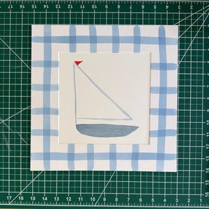 May include: A square art print featuring a minimalist sailboat illustration with a grey hull, white sail, and a small red flag. The artwork is framed by a wide border decorated with a light blue and white hand-painted plaid pattern.