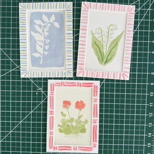 May include: Three rectangular art prints featuring botanical illustrations. One shows a white plant silhouette on a blue background with a striped border. Another displays lily of the valley in a pink striped frame. The third shows red flowers.