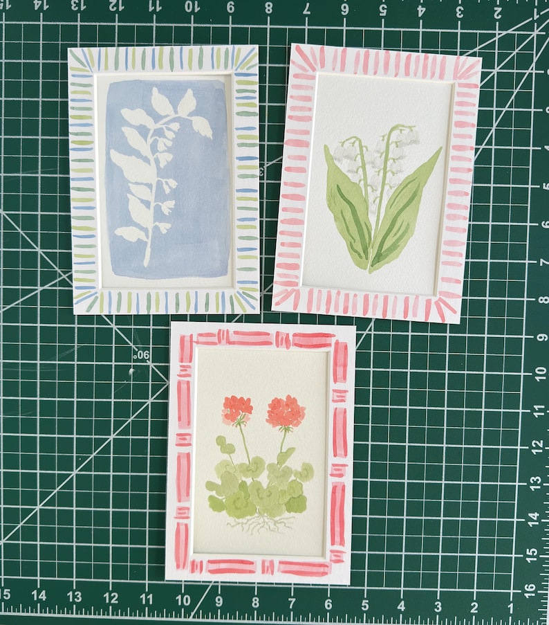May include: Three rectangular art prints featuring botanical illustrations. One shows a white plant silhouette on a blue background with a striped border. Another displays lily of the valley in a pink striped frame. The third shows red flowers.