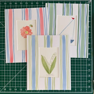 May include: Three matted art prints featuring watercolor illustrations. One print shows a coral poppy, one a blue sailboat, and one a lily of the valley. Each print is framed by a white mat with vertical stripes in coral, blue, and green tones.