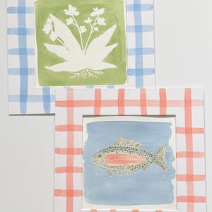 May include: Two square art prints with white borders featuring a blue and white gingham pattern on one and a coral and white gingham pattern on the other. The first print shows a white botanical illustration on a green background, while the second print displays a speckled fish with a pink stripe on a light blue background.