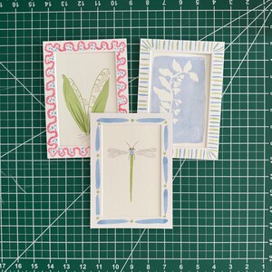 May include: Three rectangular art prints featuring hand-painted illustrations. One shows lily of the valley in a pink scalloped frame, one a blue botanical silhouette in a striped frame, and one a green dragonfly in a blue and green dotted frame.