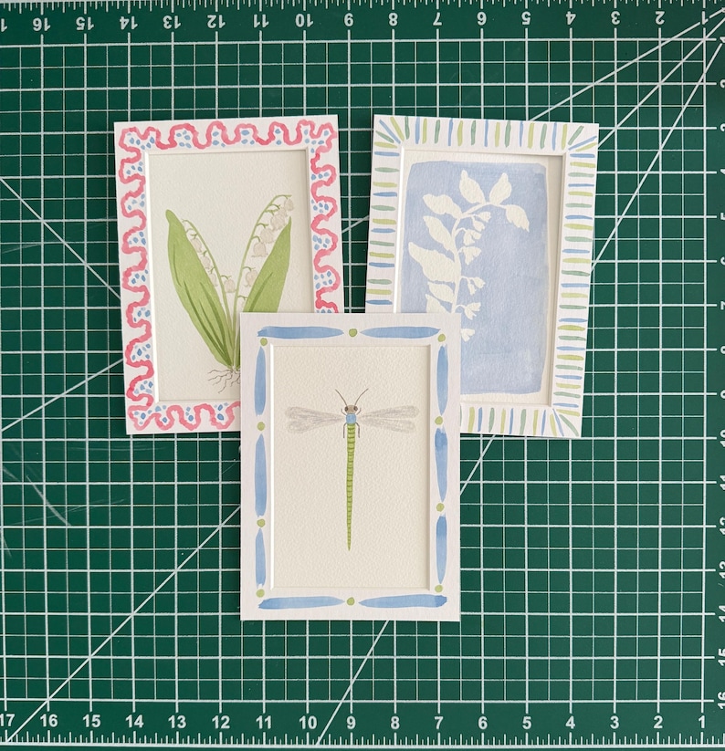 May include: Three rectangular art prints featuring hand-painted illustrations. One shows lily of the valley in a pink scalloped frame, one a blue botanical silhouette in a striped frame, and one a green dragonfly in a blue and green dotted frame.