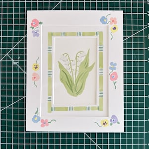 Puede incluir: Un paspart&uacute; rectangular blanco con una ilustraci&oacute;n central en acuarela de lirios de los valles blancos con hojas verdes, rodeado por un borde verde estilo bamb&uacute; y peque&ntilde;os acentos florales coloridos en las esquinas.