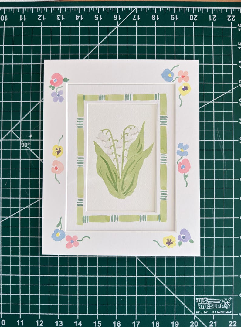 Puede incluir: Un paspart&uacute; rectangular blanco con una ilustraci&oacute;n central en acuarela de lirios de los valles blancos con hojas verdes, rodeado por un borde verde estilo bamb&uacute; y peque&ntilde;os acentos florales coloridos en las esquinas.