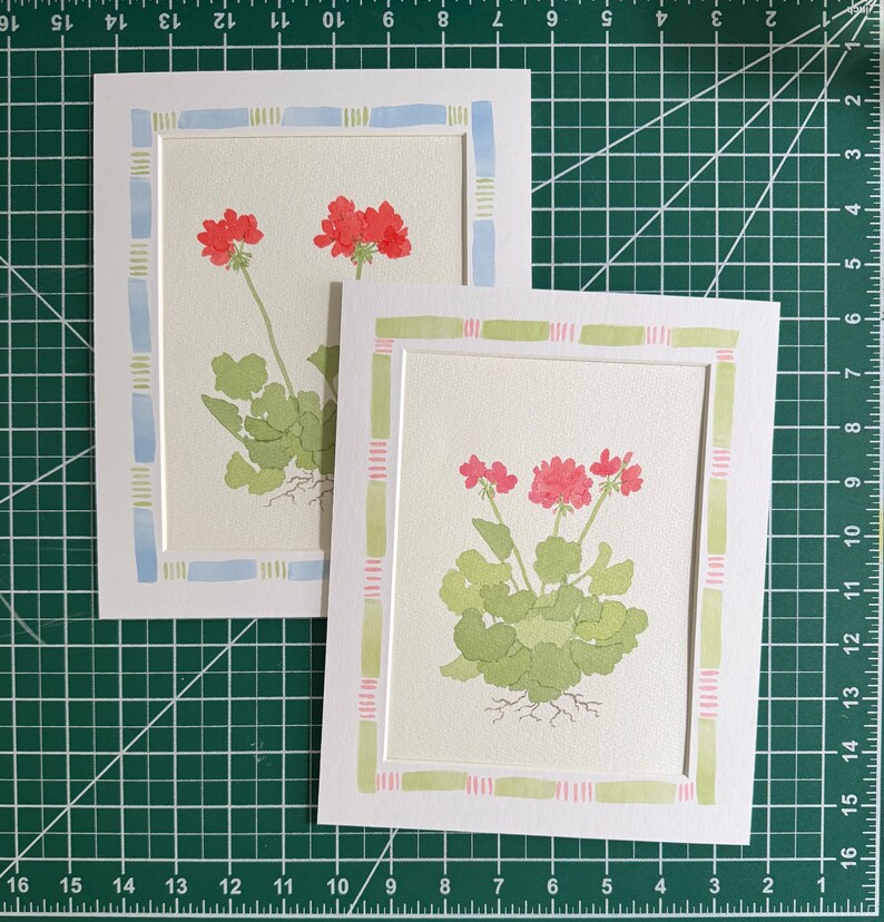 Puede incluir: Dos tarjetas de felicitaci&oacute;n rectangulares con ilustraciones en acuarela de geranios rojos con hojas verdes y ra&iacute;ces visibles. Una tarjeta tiene un borde azul claro y verde, mientras que la otra tiene un borde a rayas verde claro y rosa.