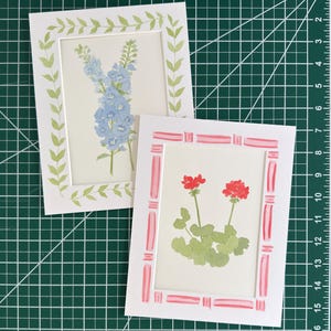 May include: Two rectangular art prints on a green cutting mat with white grid lines. One print features blue delphinium flowers with a green leaf border. The other print displays red geranium flowers with a pink and white striped border.
