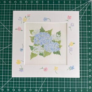 May include: A square white mat board featuring a central watercolor painting of blue and white hydrangea flowers with green leaves. The border of the mat is decorated with small, hand-painted pastel flowers in pink, yellow, and light blue hues.