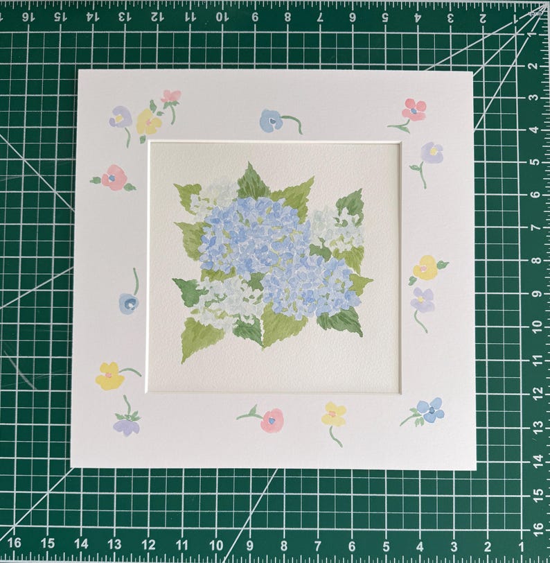 May include: A square white mat board featuring a central watercolor painting of blue and white hydrangea flowers with green leaves. The border of the mat is decorated with small, hand-painted pastel flowers in pink, yellow, and light blue hues.