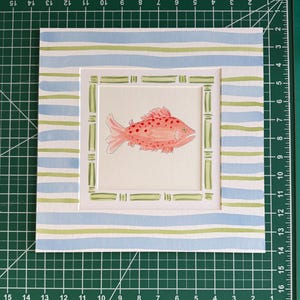 May include: A square watercolor painting of a pink fish with red spots, centered within a green bamboo-style frame. The border features horizontal light blue and green stripes. The artwork measures 12 inches by 12 inches on a cutting mat.