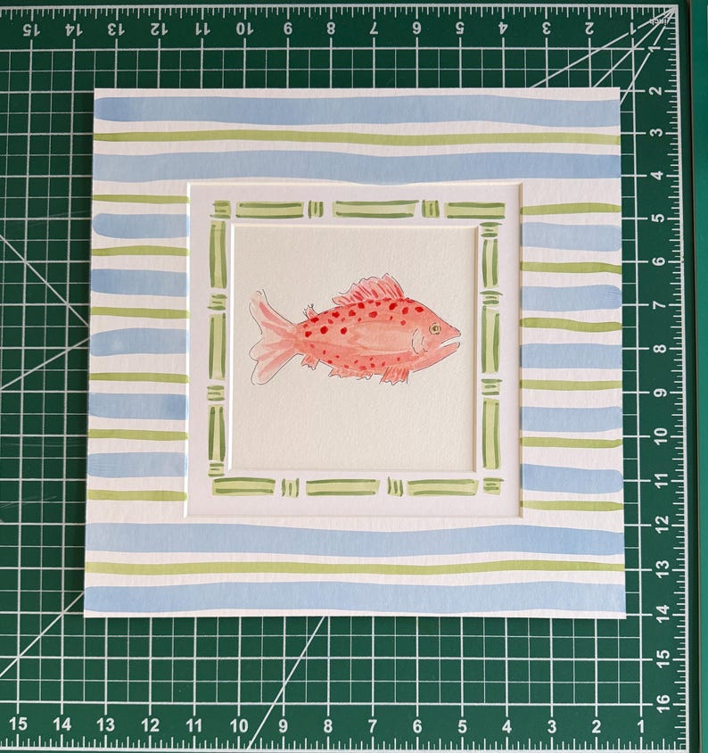 May include: A square watercolor painting of a pink fish with red spots, centered within a green bamboo-style frame. The border features horizontal light blue and green stripes. The artwork measures 12 inches by 12 inches on a cutting mat.