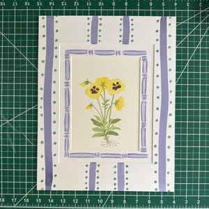 Puede incluir: Una l&aacute;mina rectangular con una ilustraci&oacute;n en acuarela de pensamientos amarillos con tallos y ra&iacute;ces verdes. La obra est&aacute; enmarcada por un borde de color violeta claro estilo bamb&uacute; y un paspart&uacute; blanco con rayas verticales.