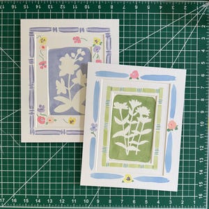 Puede incluir: Dos l&aacute;minas de arte rectangulares con siluetas florales blancas sobre fondos s&oacute;lidos. Una l&aacute;mina tiene un fondo color pervinca con un marco estilo bamb&uacute; morado, la otra un fondo verde salvia con un marco estilo bamb&uacute; verde y detalles azules.