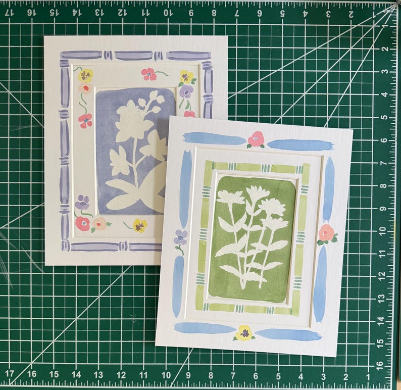 Puede incluir: Dos l&aacute;minas de arte rectangulares con siluetas florales blancas sobre fondos s&oacute;lidos. Una l&aacute;mina tiene un fondo color pervinca con un marco estilo bamb&uacute; morado, la otra un fondo verde salvia con un marco estilo bamb&uacute; verde y detalles azules.