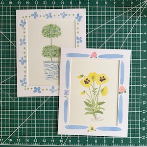 Puede incluir: Dos l&aacute;minas rectangulares sobre papel blanco. Una muestra un topiario verde de dos bolas en una maceta azul y blanca, enmarcado por flores azules y puntos verdes. La otra presenta pensamientos amarillos con tallos y ra&iacute;ces de color verde.