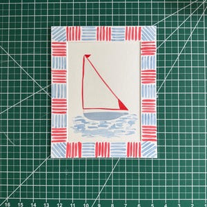 Puede incluir: Una impresi&oacute;n art&iacute;stica rectangular que presenta una ilustraci&oacute;n central de un velero con vela roja sobre agua azul claro. La obra est&aacute; rodeada por un borde decorativo de patrones de rayas horizontales y verticales en rojo y azul.