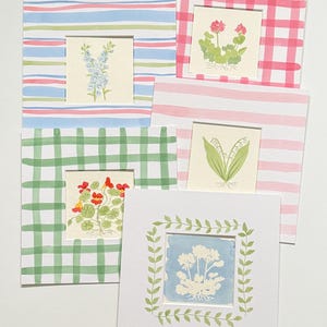 May include: A collection of five square greeting cards featuring botanical illustrations. Each card displays a different plant, including blue flowers, pink geraniums, lily of the valley, nasturtiums, and a white floral silhouette framed by green leaves.