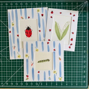 May include: Three square art prints featuring watercolor illustrations. One shows a red ladybug, one a green lily of the valley, and one a green caterpillar. The prints have decorative borders with blue brushstrokes, small insects, and yellow flowers.