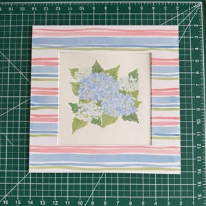 May include: A square watercolor painting of blue hydrangea flowers with green leaves, centered on a white mat board featuring a horizontal striped pattern in pink, light blue, and green, positioned on a green cutting mat with inch markings.