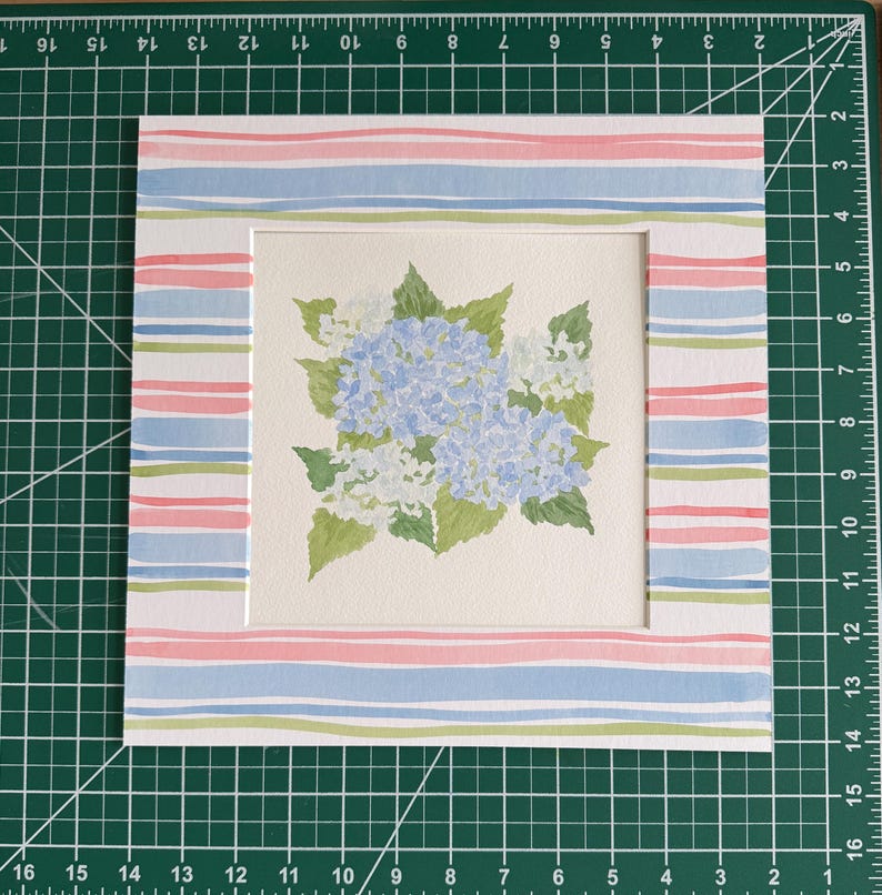 May include: A square watercolor painting of blue hydrangea flowers with green leaves, centered on a white mat board featuring a horizontal striped pattern in pink, light blue, and green, positioned on a green cutting mat with inch markings.