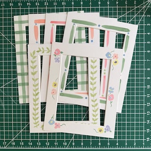Puede incluir: Un conjunto de cinco marcos rectangulares de papel blanco con diversos dise&ntilde;os pintados a mano, que incluyen patrones de cuadros verdes, rayas verticales rosas y verdes, y delicados motivos florales en tonos pastel de rosa, azul y amarillo.