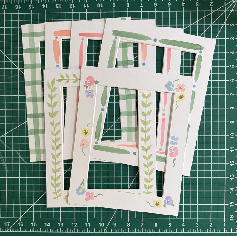 Puede incluir: Un conjunto de cinco marcos rectangulares de papel blanco con diversos dise&ntilde;os pintados a mano, que incluyen patrones de cuadros verdes, rayas verticales rosas y verdes, y delicados motivos florales en tonos pastel de rosa, azul y amarillo.