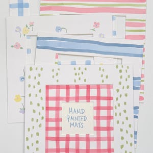 May include: A collection of five rectangular paper picture mats featuring various hand-painted designs including blue plaid, pink and green stripes, small floral patterns, blue horizontal stripes, and a red plaid border with the text HAND PAINTED MATS.