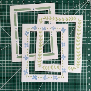 Puede incluir: Tres marcos rectangulares de papel blanco con dise&ntilde;os pintados a mano. Un marco presenta pinceladas verdes y puntos azules. Otro tiene un patr&oacute;n de enredadera verde. El tercero muestra un borde de flores azules y peque&ntilde;os puntos verdes.