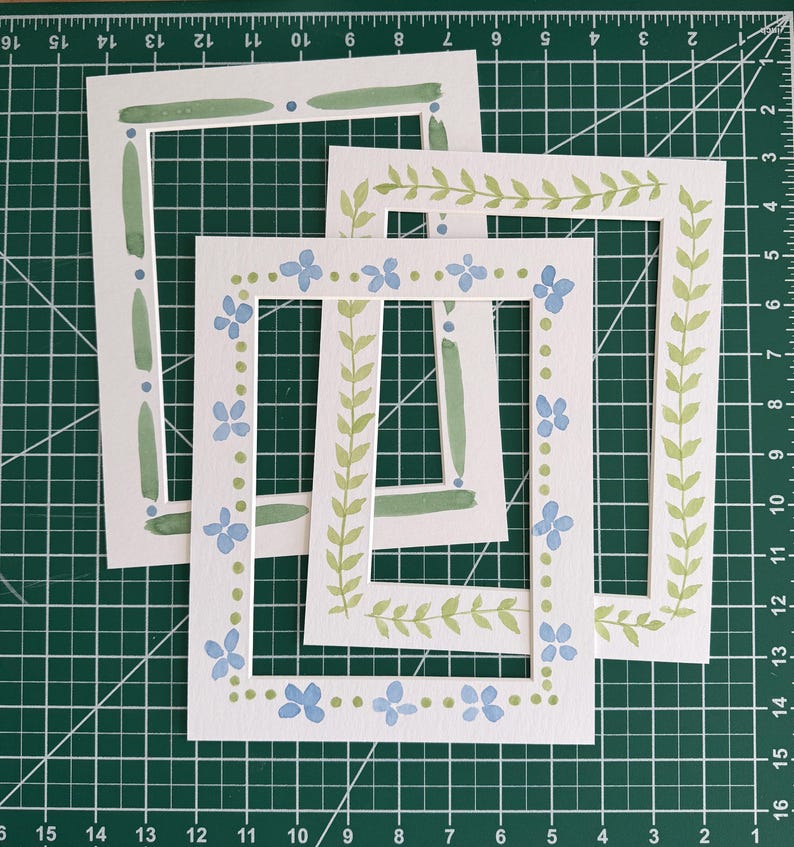 Puede incluir: Tres marcos rectangulares de papel blanco con dise&ntilde;os pintados a mano. Un marco presenta pinceladas verdes y puntos azules. Otro tiene un patr&oacute;n de enredadera verde. El tercero muestra un borde de flores azules y peque&ntilde;os puntos verdes.