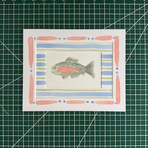 Puede incluir: Una pintura rectangular de acuarela que muestra un pez moteado con una franja lateral rosa, centrado sobre un fondo de rayas horizontales azul claro y verde p&aacute;lido, enmarcado por un paspart&uacute; blanco con pinceladas rosas y puntos azules.