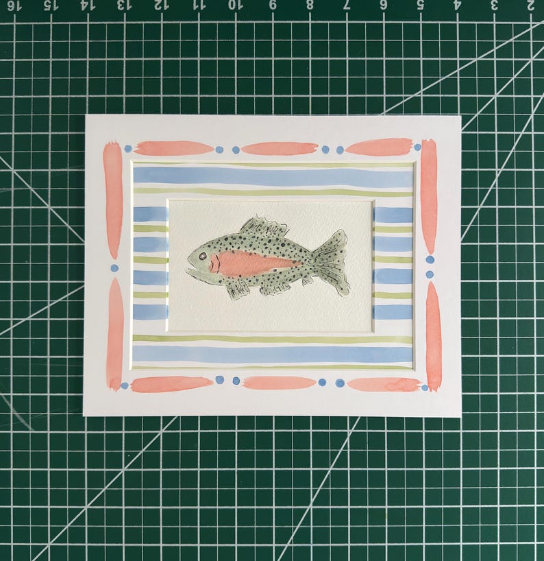 Puede incluir: Una pintura rectangular de acuarela que muestra un pez moteado con una franja lateral rosa, centrado sobre un fondo de rayas horizontales azul claro y verde p&aacute;lido, enmarcado por un paspart&uacute; blanco con pinceladas rosas y puntos azules.