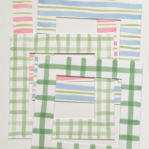 May include: A collection of four rectangular paper picture frame mats featuring hand-painted watercolor patterns. The designs include horizontal pink stripes, blue and green stripes, and green checkered patterns on a white background.
