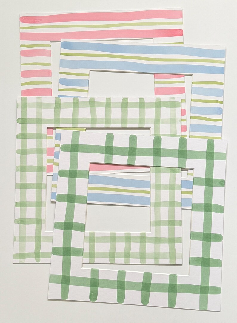 May include: A collection of four rectangular paper picture frame mats featuring hand-painted watercolor patterns. The designs include horizontal pink stripes, blue and green stripes, and green checkered patterns on a white background.