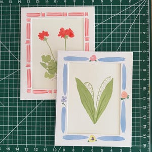 May include: Two rectangular art prints on white matting. One print features red geraniums with green leaves and a pink striped border. The other print displays white lily of the valley flowers with green leaves, a blue brushstroke border, and small floral accents.