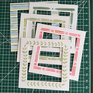 May include: A collection of square paper frames with watercolor designs. The frames feature various patterns, including stripes, leaves, and geometric shapes in shades of green, pink, and blue. The frames are arranged on a green cutting mat.