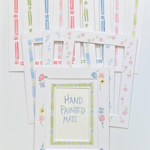 Puede incluir: Una colecci&oacute;n de paspart&uacute;s rectangulares blancos para marcos de fotos con dise&ntilde;os pintados a mano. Los paspart&uacute;s muestran varios patrones, incluyendo rayas coloridas, bordes estilo bamb&uacute; y motivos florales. Uno lleva el texto Hand Painted Mats.