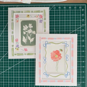 Puede incluir: Dos l&aacute;minas de arte rectangulares con bordes pintados a mano. La l&aacute;mina izquierda presenta un fondo verde salvia con siluetas florales blancas y un borde de rayas verdes. La derecha muestra una amapola roja con un borde de lazos rosas y azules.