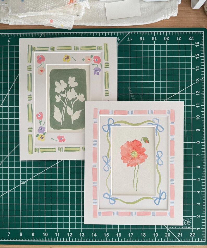 Puede incluir: Dos l&aacute;minas de arte rectangulares con bordes pintados a mano. La l&aacute;mina izquierda presenta un fondo verde salvia con siluetas florales blancas y un borde de rayas verdes. La derecha muestra una amapola roja con un borde de lazos rosas y azules.