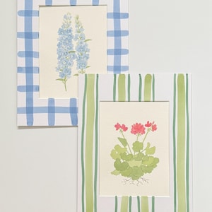 May include: Two rectangular art prints featuring botanical illustrations. One print displays blue delphinium flowers framed by a blue and white gingham border. The second print shows a pink geranium plant with green leaves inside a green striped border.