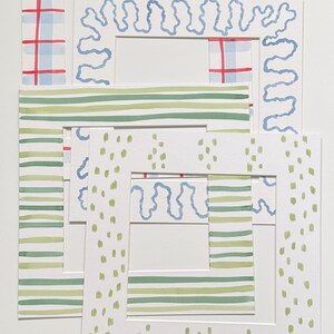 May include: A set of four square paper picture mats featuring hand-painted designs. The patterns include a blue and red plaid, a light blue wavy line, horizontal green stripes, and light green vertical dashes on a white background.