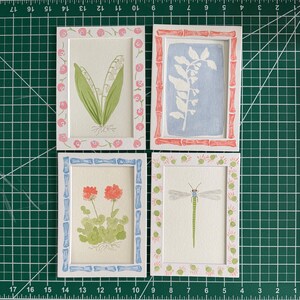 May include: Four rectangular white frames containing watercolor illustrations. Top left: lily of the valley with a pink floral border. Top right: white botanical silhouette on blue with a red bamboo-style border. Bottom left: orange flowers with a blue bamboo-style border. Bottom right: green dragonfly with a pink and green dotted border.